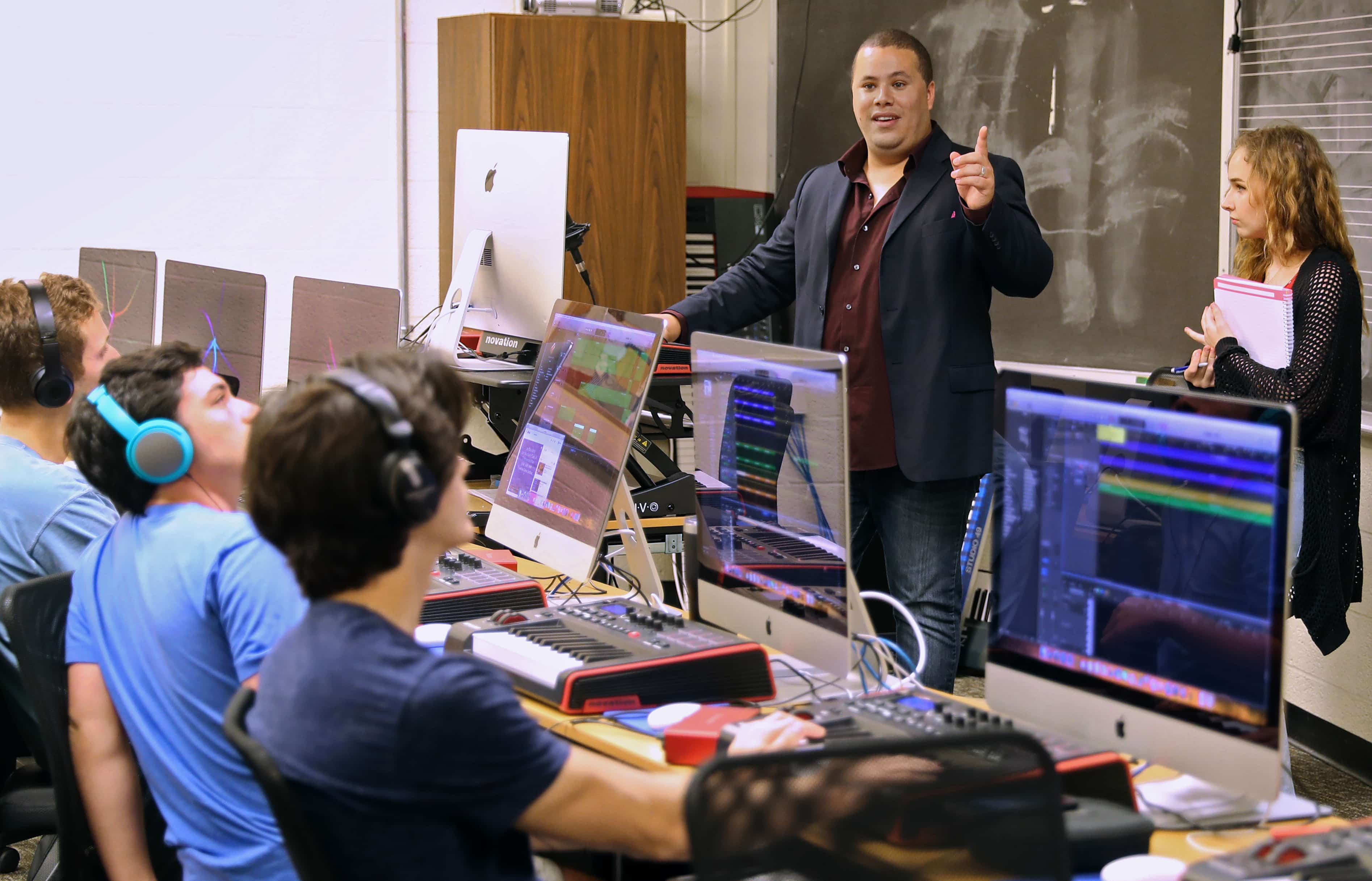 Professor Jose Valentino Ruiz in a music technology classroom talking to students who are working on iMac computers