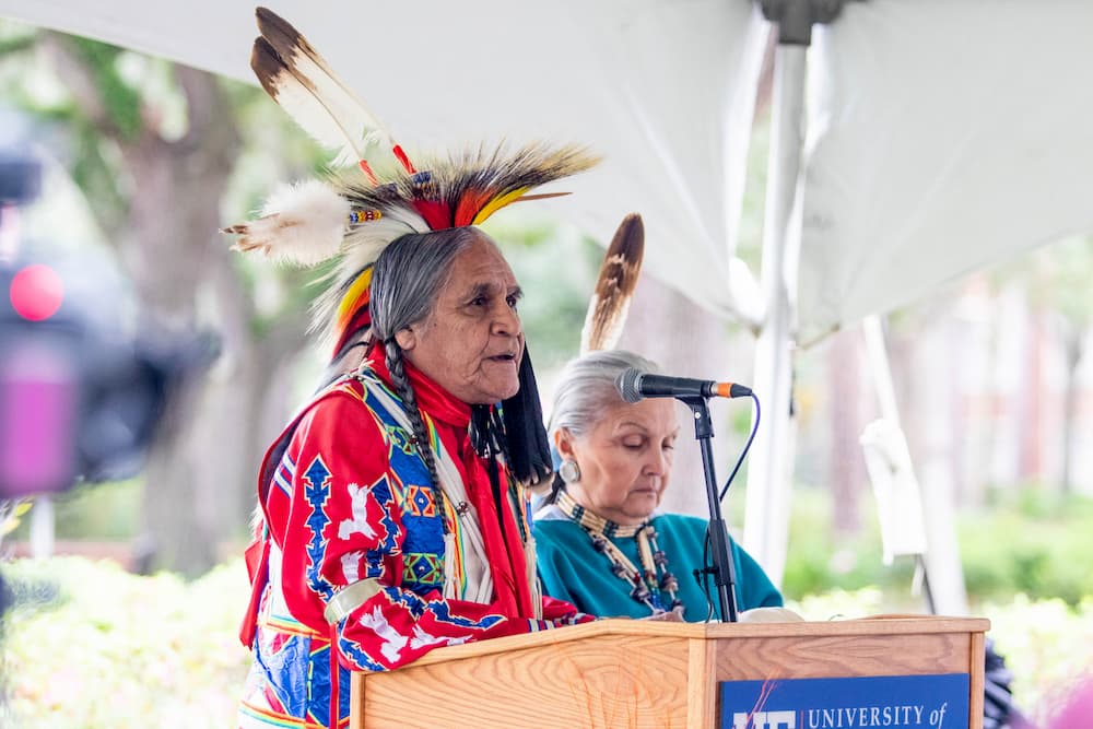 Man wearing traditional Kiowa clothing speaks at a lectern. 